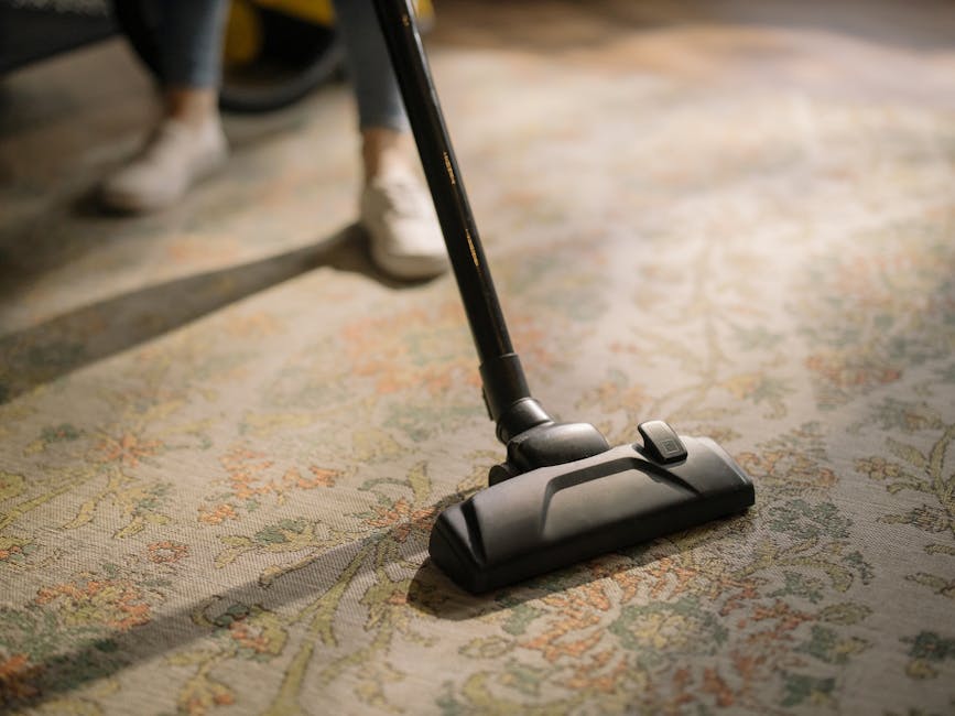 A man with gray hair and a beard is performing domestic surface cleaning in a bright living room, using a vacuum cleaner. The room features a light-colored sofa adorned with orange and blue cushions,