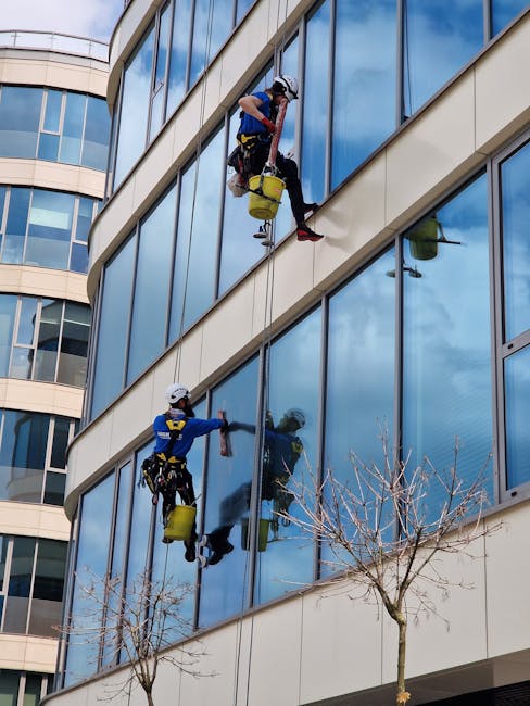 Two professional window cleaners wearing safety harnesses and helmets are performing surface cleaning on the exterior glass windows of a modern multi-storey office building in daylight. They use high-
