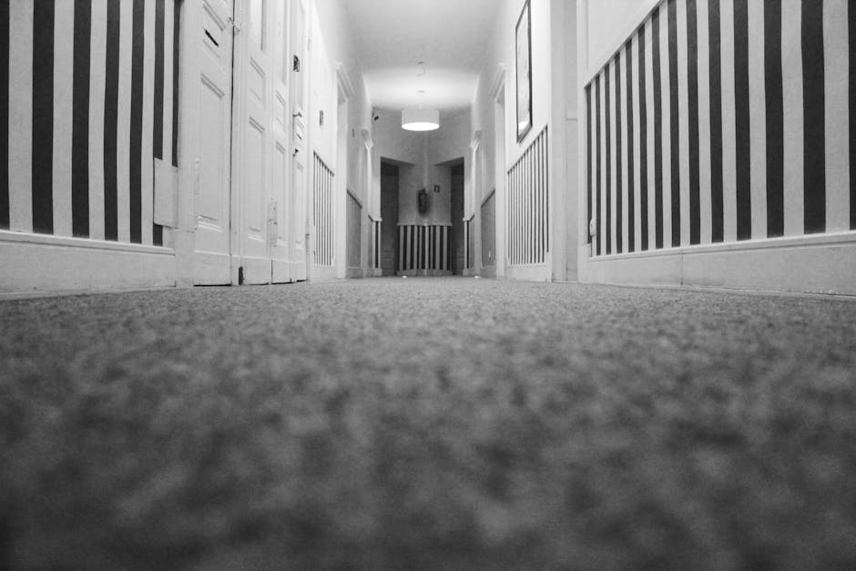 A low-angle view of a clean, soft-textured carpeted corridor in a residential or hotel setting, with white wooden door panels and striped wallpaper on the walls. The corridor features a bright ceiling