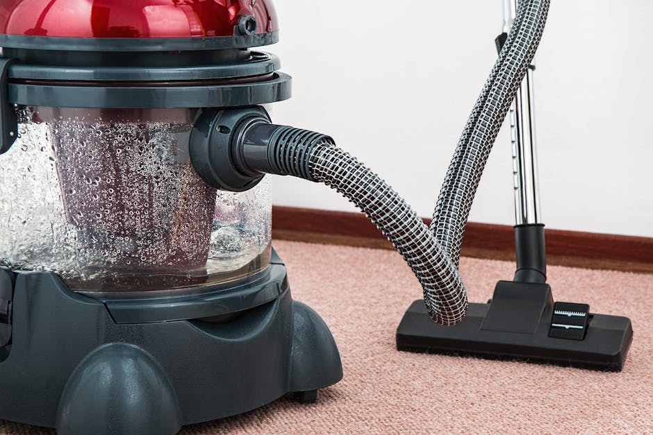 Close-up of a modern vacuum cleaner with a red top and clear dust container, positioned on a pink carpeted floor in a room with white walls and wooden skirting. The vacuum's flexible hose, covered in a black and white patterned fabric, extends towards the floor, with the hose resting on a black floor attachment. The scene highlights the surface cleaning process used by Hounslow Carpet Cleaners, showcasing deep cleaning equipment used for maintaining hygienic and spotless domestic and commercial carpets, aligning with the services offered on the Trusted carpet cleaning team Hounslow High Street local webpage.